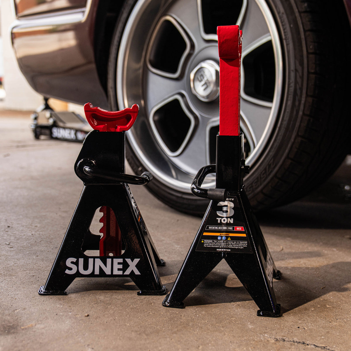 A pair of SUNEX Jack Stands placed just in front of a truck and its rear wheel with a floor jack shown in the rear.