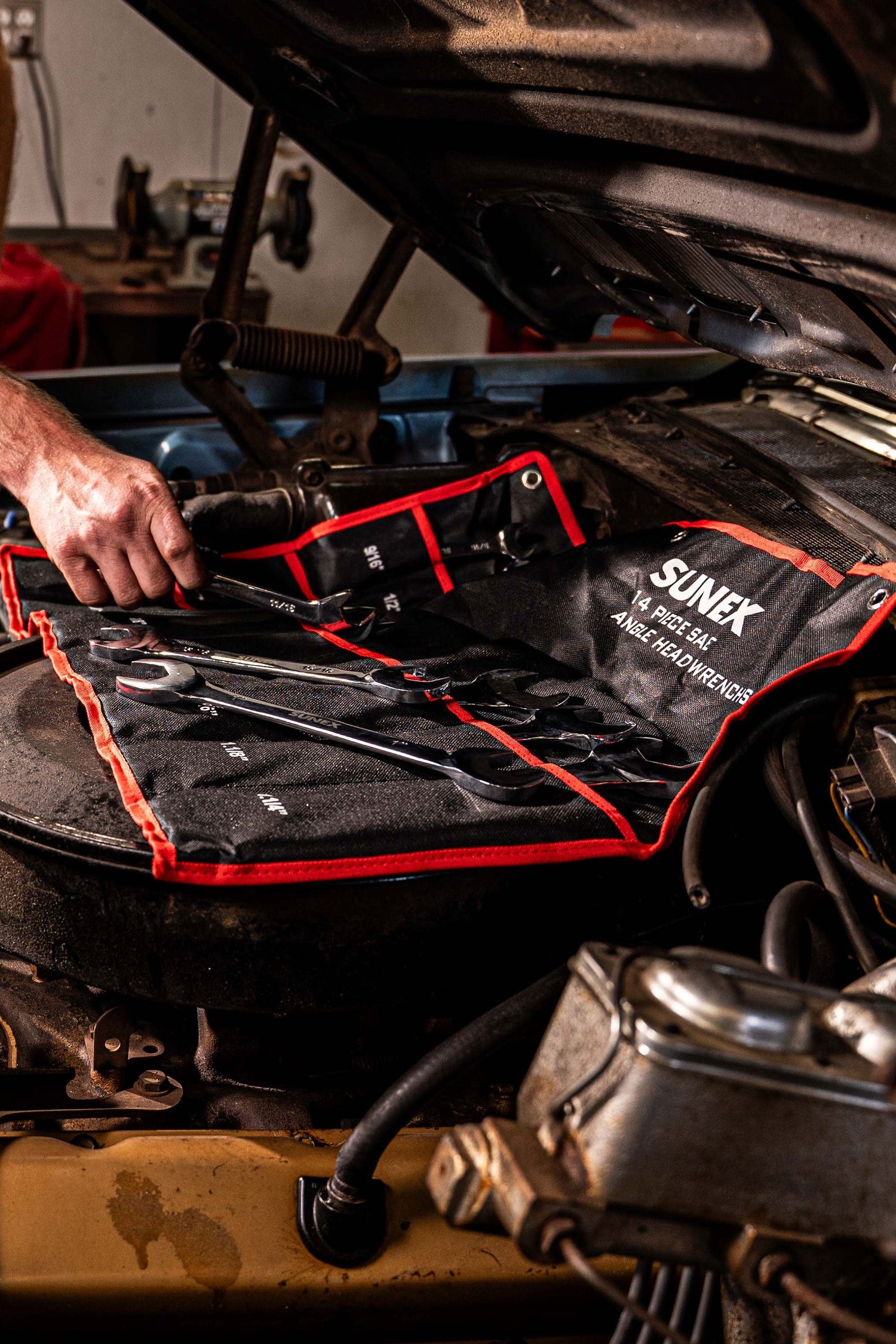 A mechanic reaches for a angle head wrench from the SUNEX 9914A SAE Angle Head Wrench Set while it is resting in and on it's tool roll inside an engine compartment of an older vehicle