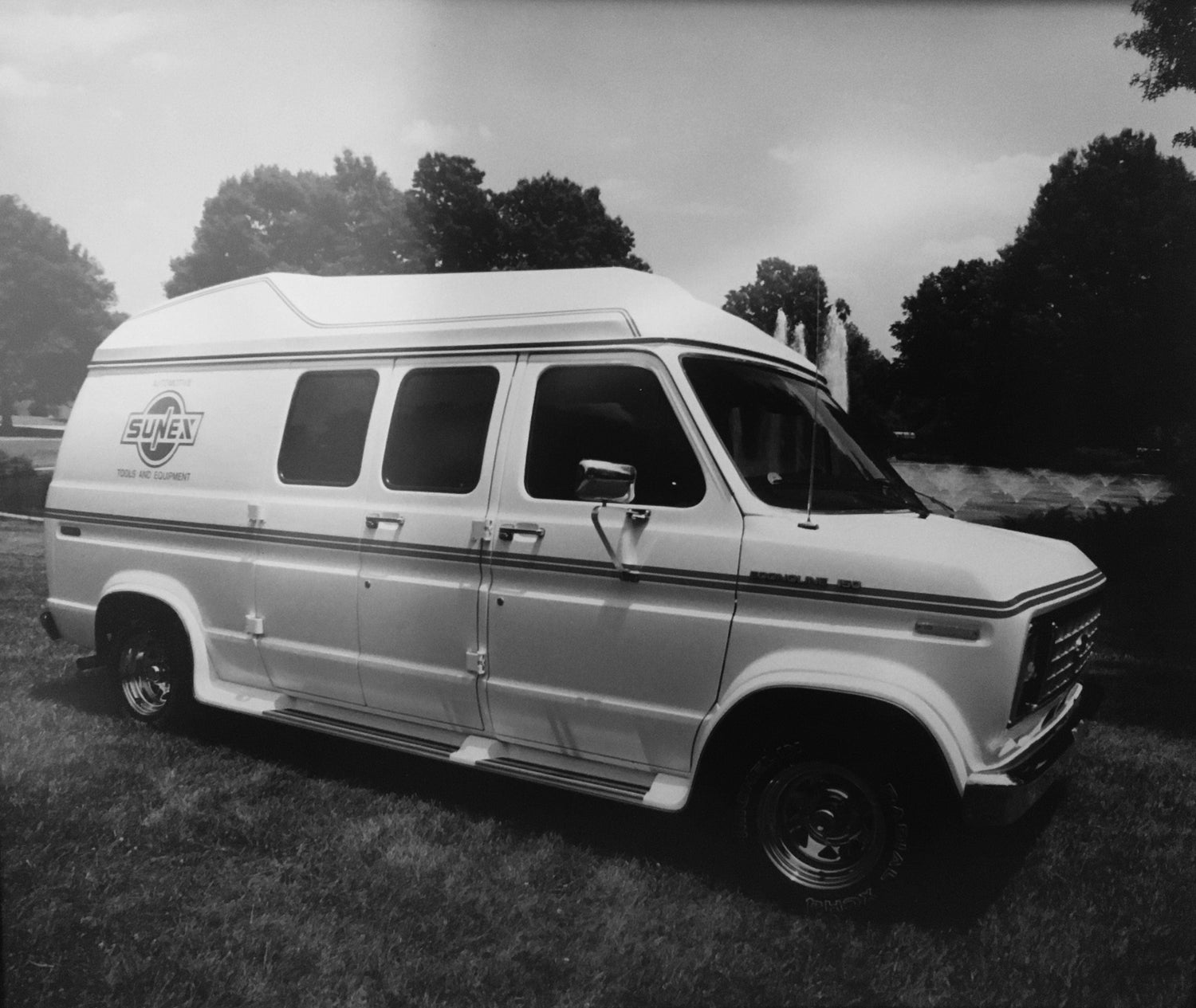 Black and White photo of SUNEX Automotive Tools and Equipment Van from 1980s on grass lawn in front of fountains.